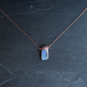 May include: A necklace featuring a raw, rectangular-shaped opal pendant with iridescent blue and white flashes. The stone is secured by a delicate copper wire wrap and hangs from a thin, matching copper-toned metal chain link necklace.
