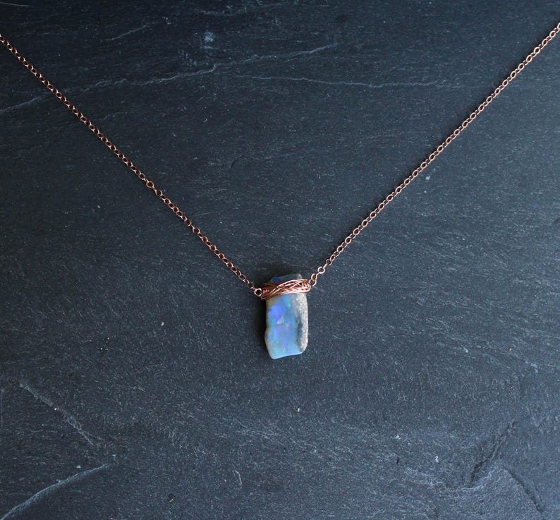 May include: A necklace featuring a raw, rectangular-shaped opal pendant with iridescent blue and white flashes. The stone is secured by a delicate copper wire wrap and hangs from a thin, matching copper-toned metal chain link necklace.