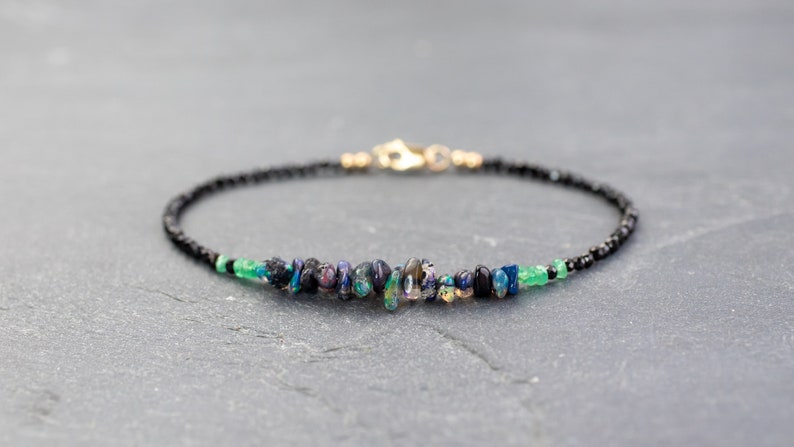 Puede incluir: Una pulsera delicada que presenta una hilera de peque&ntilde;as cuentas negras, acentuada con una fila central de fragmentos de &oacute;palo iridiscente y peque&ntilde;as cuentas de piedras preciosas verdes. La pieza incluye un cierre de mosquet&oacute;n de metal dorado.
