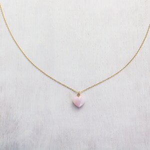 May include: A delicate gold chain necklace featuring a small, smooth, light pink heart-shaped pendant. The pendant is suspended from a thin, polished gold-tone metal chain that forms a gentle curve, showcasing a simple and refined jewelry design.
