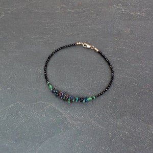 Puede incluir: Una pulsera delicada que presenta una hilera de peque&ntilde;as cuentas negras facetadas, acentuada con un grupo de piedras irregulares iridiscentes en tonos verde, azul y morado, terminada con un cierre de mosquet&oacute;n de metal en tono dorado.