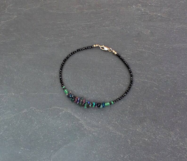 Puede incluir: Una pulsera delicada que presenta una hilera de peque&ntilde;as cuentas negras facetadas, acentuada con un grupo de piedras irregulares iridiscentes en tonos verde, azul y morado, terminada con un cierre de mosquet&oacute;n de metal en tono dorado.