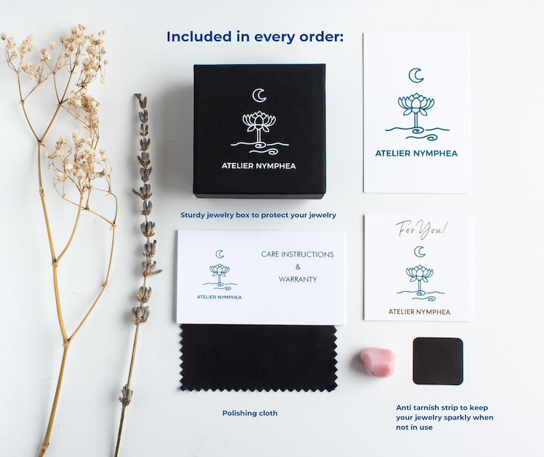 K&ouml;nnte beinhalten: Eine schwarze quadratische Schmuckschatulle mit wei&szlig;em Lotus- und Mondlogo, eine wei&szlig;e Karte mit demselben Logo und dem Text ATELIER NYMPHEA, eine Pflegeanleitung, ein schwarzes Poliertuch, ein kleiner rosa Stein und ein schwarzer Anlaufschutzstreifen.