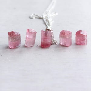 K&ouml;nnte beinhalten: Eine Silberkettenhalskette mit einem Anh&auml;nger aus rohem, durchscheinendem rosa Turmalinkristall, begleitet von vier weiteren losen, nicht gefassten rohen rosa Turmalinkristallen in verschiedenen Formen und Gr&ouml;&szlig;en in einer horizontalen Reihe.