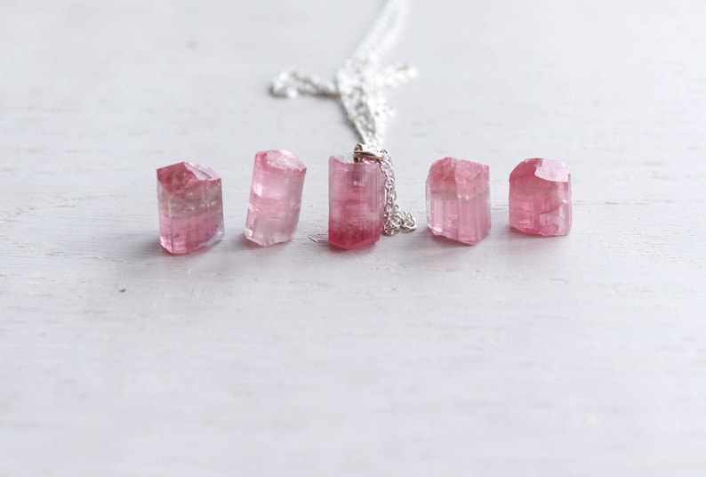 K&ouml;nnte beinhalten: Eine Silberkettenhalskette mit einem Anh&auml;nger aus rohem, durchscheinendem rosa Turmalinkristall, begleitet von vier weiteren losen, nicht gefassten rohen rosa Turmalinkristallen in verschiedenen Formen und Gr&ouml;&szlig;en in einer horizontalen Reihe.