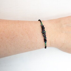 Puede incluir: Una pulsera que presenta una serie de peque&ntilde;as piedras preciosas irregulares y pulidas en tonos morado intenso, azul oscuro y verde vibrante, ensartadas en un cord&oacute;n fino y oscuro que rodea la mu&ntilde;eca con un aspecto delicado y sencillo.