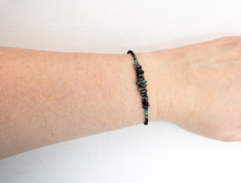 Puede incluir: Una pulsera que presenta una serie de peque&ntilde;as piedras preciosas irregulares y pulidas en tonos morado intenso, azul oscuro y verde vibrante, ensartadas en un cord&oacute;n fino y oscuro que rodea la mu&ntilde;eca con un aspecto delicado y sencillo.