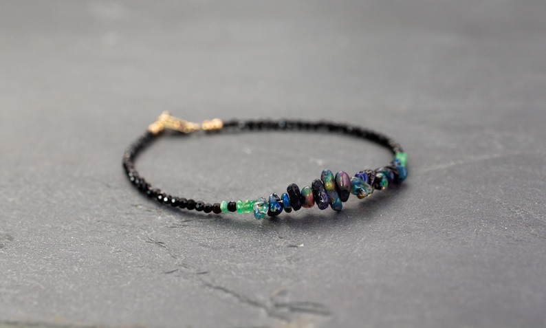 Puede incluir: Una pulsera delicada con una hilera de peque&ntilde;as cuentas negras facetadas y un grupo central de piedras oscuras irregulares y pulidas con destellos iridiscentes en azul, verde y morado. La pieza incluye un peque&ntilde;o cierre de metal dorado.