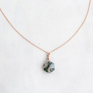 May include: A delicate rose gold chain necklace featuring a hexagonal moss agate pendant. The pendant is suspended from a small rose gold bead and a jump ring, showcasing the translucent stone with its distinct green moss-like inclusions.