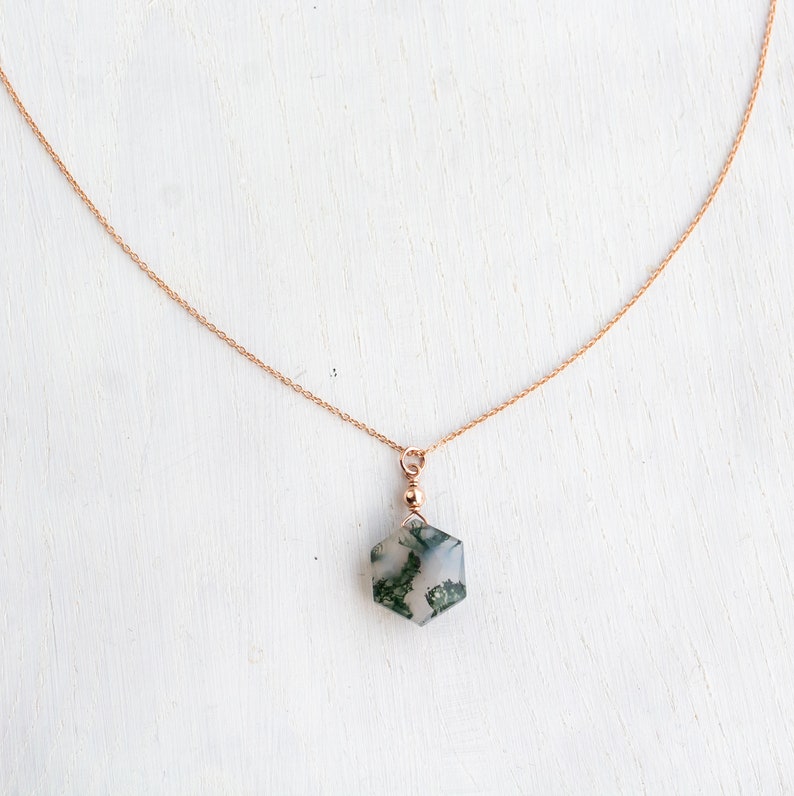 May include: A delicate rose gold chain necklace featuring a hexagonal moss agate pendant. The pendant is suspended from a small rose gold bead and a jump ring, showcasing the translucent stone with its distinct green moss-like inclusions.
