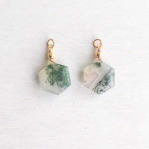 May include: Two hexagonal moss agate pendants featuring translucent white stone with green dendritic inclusions. Each pendant is topped with a small gold-toned metal bead and a circular jump ring for attachment, showcasing natural stone patterns.