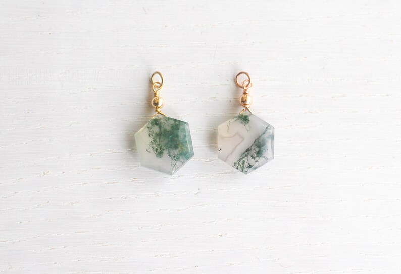 May include: Two hexagonal moss agate pendants featuring translucent white stone with green dendritic inclusions. Each pendant is topped with a small gold-toned metal bead and a circular jump ring for attachment, showcasing natural stone patterns.