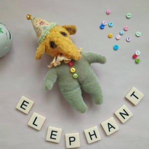 May include: A yellow stuffed elephant wearing a green and white outfit with a party hat. The elephant is surrounded by colorful buttons and the word "ELEPHANT" spelled out in letter tiles.