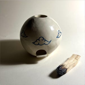 Può includere: Bruciatore di incenso in ceramica bianca di forma rotonda con disegni di nuvole blu. È incluso un bastoncino di incenso in legno.