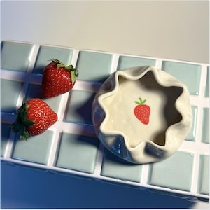 Könnte beinhalten: Eine weiße Keramikschale mit einem gewellten Rand und einem kleinen roten Erdbeerdesign. Zwei rote Erdbeeren liegen auf einer hellblauen gekachelten Oberfläche.