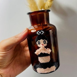 Puede incluir: Una botella de vidrio marrón con una pegatina de muñeca de cuentas en la parte delantera. La muñeca lleva un traje de baño negro y tiene el pelo negro y los ojos blancos. La botella está llena de hierba de la pampa seca.