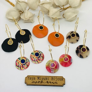 Könnte beinhalten: Eine Auswahl an bunten Creolen mit verschiedenen Designs. Jeder Ohrring hat einen runden Anhänger mit einem kleinen Edelstein in der Mitte. Die Anhänger sind aus verschiedenen Materialien gefertigt, darunter lackiertes Metall, Harz und Holz. Die Ohrringe werden auf einer weißen Oberfläche mit einem Holzschild präsentiert, auf dem "Yaya Miyuki Bijoux" steht.