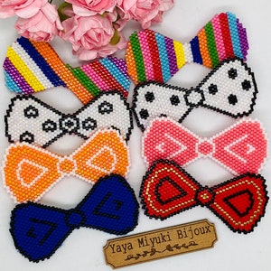 Può includere: Una collezione di spille a farfalla fatte a mano con perline in vari colori e motivi, tra cui strisce arcobaleno, pois e colori solidi. Le spille sono esposte su una superficie bianca con un'etichetta marrone che recita "Yaya Miyuki Bijoux".