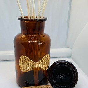 May include: A brown glass bottle with a gold bow design and a wooden label that reads "Yaya Miyuki Bijoux". The bottle is filled with wooden sticks and has a black lid with the text "250ml" printed on it.