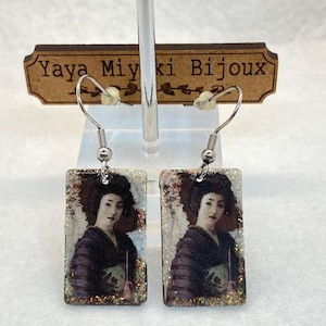 May include: Rectangular earrings with a portrait of a woman in traditional Japanese dress. The earrings hang from silver hooks and are decorated with glitter. The brand name "Yaya Miyki Bijoux" is displayed on a wooden sign. A pair of earrings.