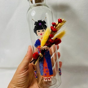Può includere: Una bottiglia di vetro trasparente con un tappo di sughero, che contiene una figura di perline di una donna con un vestito blu e arancione, che tiene un mazzo di fiori secchi.