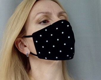 Może przedstawiać: Czarna maska ​​na twarz z białymi kropkami. Maska ​​jest wykonana z tkaniny i ma elastyczne sznurki do uszu.