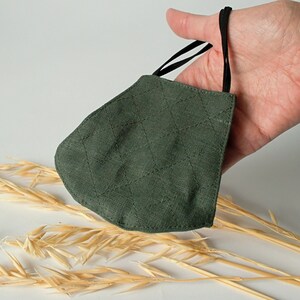 Può includere: Una maschera facciale in tessuto trapuntato verde scuro con lacci elastici neri. La maschera è appoggiata su una superficie con steli di grano secchi.
