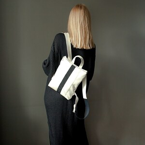 May include: A white backpack with a black vertical stripe and white straps. The bag has a top handle and is worn over the shoulders. The person in the image is wearing a black dress.