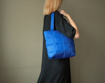 Könnte beinhalten: Eine leuchtend blaue, gesteppte Tragetasche mit einem Gittermuster und einem einzelnen Schulterriemen. Die Tasche zeichnet sich durch eine weiche, gepolsterte Textur und eine rechteckige Form aus, die einen lebendigen Kontrast zur dunklen Kleidung bildet.