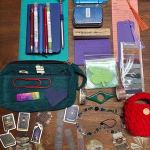May include: A collection of stationery items including a teal corduroy pencil case, various pens, a blue notebook, a reading list card with a tassel, a purple bookmark, sticky notes, washi tape, a beaded charm, and assorted book-themed stickers.