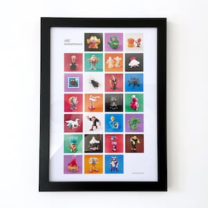 Op de afbeelding: Een ingelijste poster met de titel "ABC Monstrueux" en 25 kleurrijke vierkanten, elk met een ander cartoonmonster. De poster is gedrukt op wit papier en heeft een zwarte lijst.