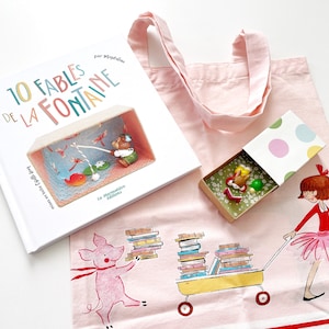 Könnte beinhalten: Eine rosa Tragetasche mit einem weißen Buch mit dem Titel "10 Fables de la Fontaine" und einer kleinen Schachtel mit buntem Punkte-Design. Die Tragetasche zeigt außerdem ein rosa Schwein, das einen Stapel Bücher trägt, und ein Mädchen in einem roten Kleid und rosa Tutu, das einen Wagen voller Bücher schiebt.
