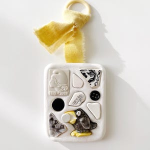 May include: A white rectangular ornament with embedded ceramic pieces, including a black and white patterned fragment, a black button, and a small bird figure. A yellow ribbon and wooden ring are attached at the top.