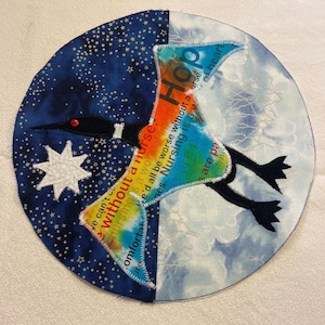 May include: A round, fabric art piece depicting a bird in flight, split into two halves. One side shows a starry night sky with a white star, the other a cloudy sky. The bird is colorful with text that reads "Hop" and "Nursing is a work of heart."