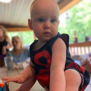 May include: A baby wearing a red and black sleeveless shirt with a large red circle design. The baby is sitting on a wooden surface and looking to the side.