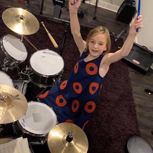 May include: A young person wearing a blue and orange dress with a donut pattern is playing a drum set. The person is holding drumsticks in each hand and is smiling. The drum set is on a dark carpet.