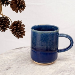 Puede incluir: Una taza de cerámica azul con un esmalte azul claro. La taza tiene un fondo redondeado y un asa.