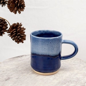 May include: A blue ceramic mug with a white glaze on the top half. The mug has a rounded bottom and a handle.