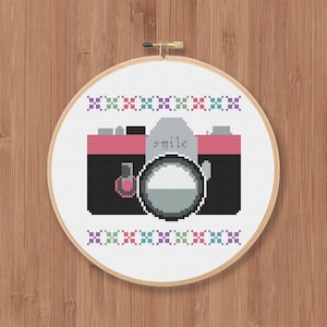 May include: A cross-stitch embroidery hoop with a black and pink camera design. The camera lens is gray and the text "smile" is written on the camera.