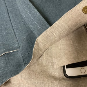 May include: A light blue linen tote bag with a beige linen lining. The bag is open and shows a black phone case with a white phone inside, and a pair of glasses. The bag has a brass button closure.