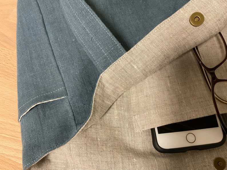 May include: A light blue linen tote bag with a beige linen lining. The bag is open and shows a black phone case with a white phone inside, and a pair of glasses. The bag has a brass button closure.