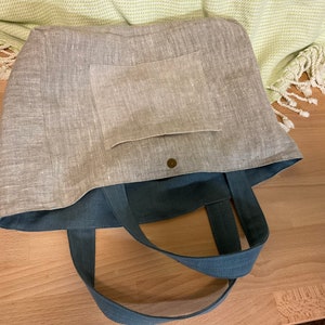 May include: A gray linen tote bag with a blue canvas lining and a small pocket on the front. The bag has two long blue canvas straps.