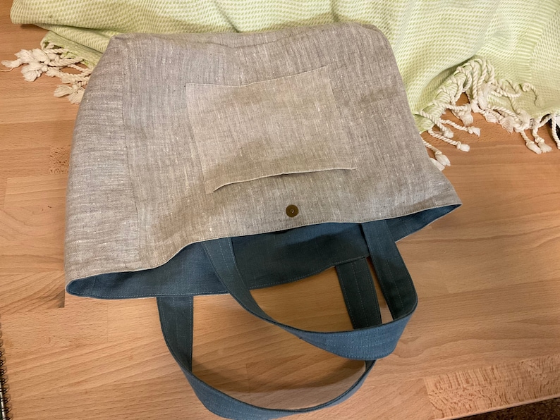 May include: A gray linen tote bag with a blue canvas lining and a small pocket on the front. The bag has two long blue canvas straps.