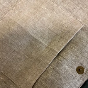 May include: Close-up of a beige linen fabric with a large pocket and a small metal button.