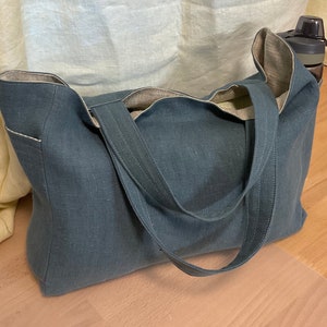 May include: A large, dark blue tote bag made of linen with a contrasting light beige lining. The bag has two long straps and a pocket on the front.
