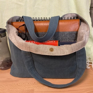 May include: A blue canvas tote bag with a brown leather notebook, a book, and a water bottle inside. The bag has a light brown linen lining.