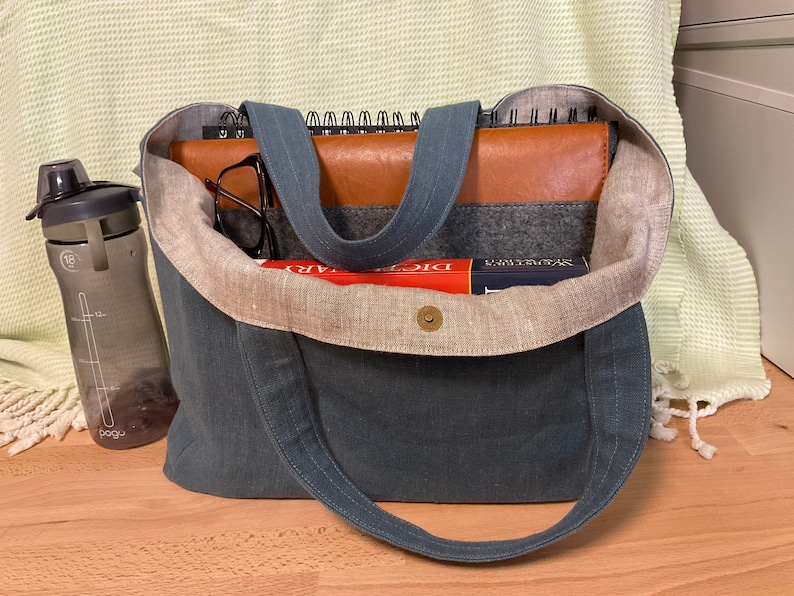 May include: A blue canvas tote bag with a brown leather notebook, a book, and a water bottle inside. The bag has a light brown linen lining.