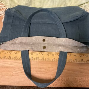 May include: A blue canvas tote bag with brown fabric lining and two brown button closures. The bag has two long straps and is measured with a ruler.