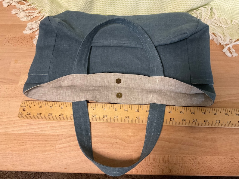 May include: A blue canvas tote bag with brown fabric lining and two brown button closures. The bag has two long straps and is measured with a ruler.