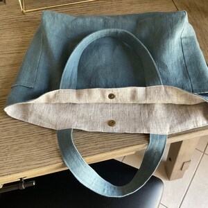 May include: A blue linen tote bag with two brown metal snaps and a light brown linen lining. The bag has two long blue linen straps.